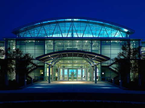An outdoor photograph of the Herman Miller Greenhouse factory at night, illuminated from inside.
