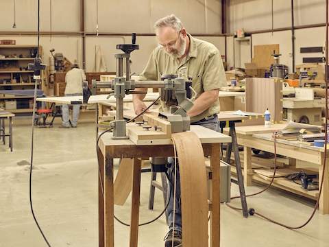 A man is examining and working on a long piece of wood veneer at a standing-height table within a large factory space.