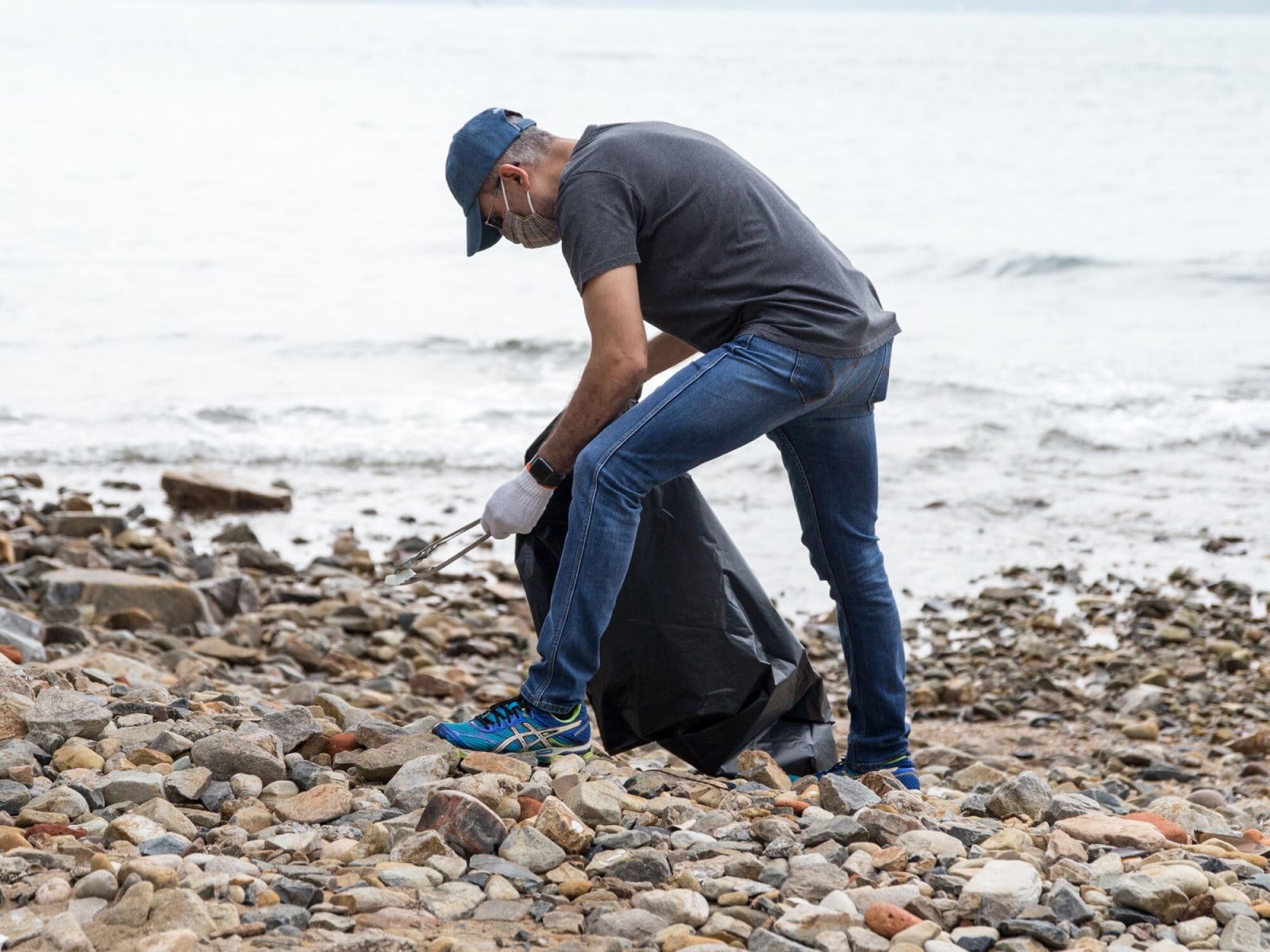 A man holding a black garbage bag is bending over to collect plastic trash on a beach.