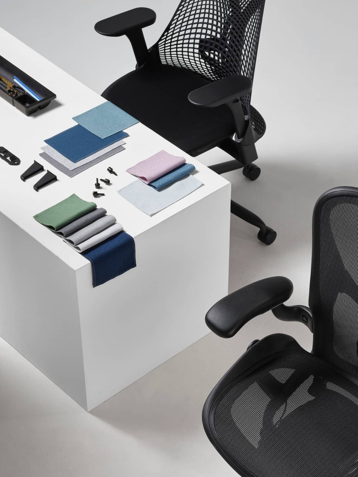 A black Aeron and Sayl office chair viewed from above, are positioned next to a white display cube, on top of which an arrangement of folded Revenio textile swatches, OE1 trolly plastic clips, and a Tu Pedestal Utility Tray made from ocean-bound plastics are all displayed.