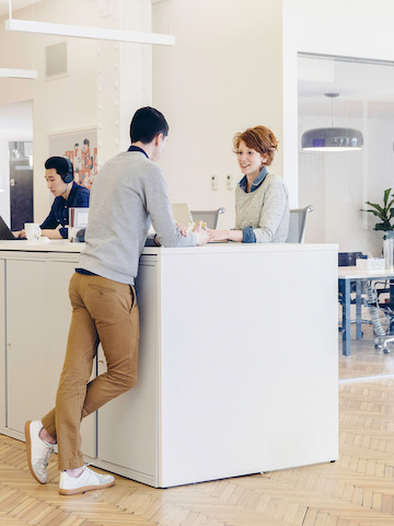 Two people talking at a standing-height work surface in the middle of an office setting.