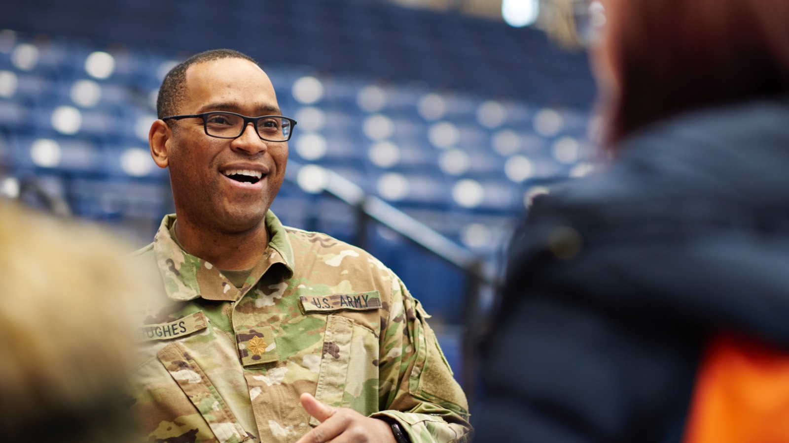 A man wearing a military uniform is smiling and interacting with people who are only partially visible within the photo.