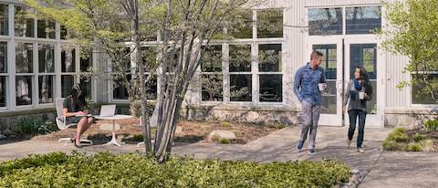 A young woman and man walk through a courtyard outside, chatting and drinking coffee while a woman works on a laptop nearby.