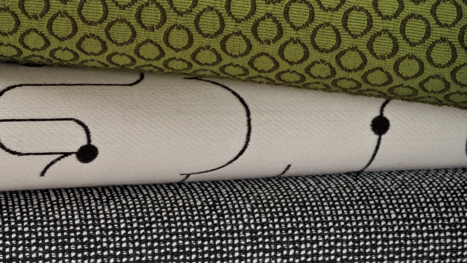Layers of folded fabric samples, including a green patterned fabric, a black and white checked fabric, and a fabric with a lined pattern.