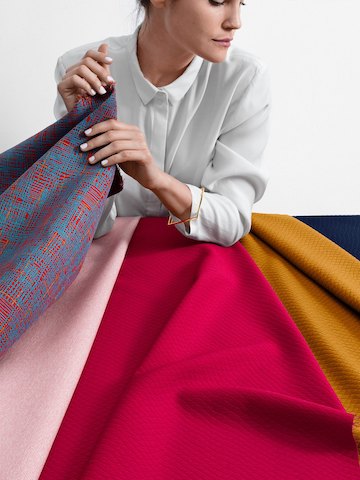 A woman wearing a white button-up shirt examines a variety of different fabric samples in a range of textures, patterns, and weaves.