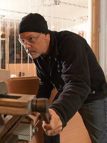 Designer Brian Alexander operando uma máquina em seu estúdio de trabalho. Selecione para ir para a página da história do sistema de mesas com regulagem de altura Renew Link.