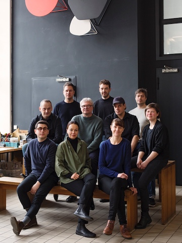 De mannen en vrouwen van Studio 7.5 zitten gegroepeerd op houten banken in hun werkruimte.