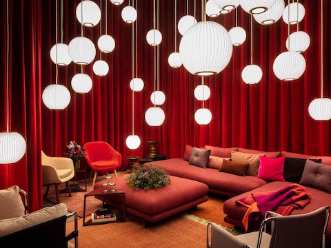 A lounge space containing a large, red Lecco Open Sectional with an ottoman; a multitude of Nelson Bubble Lamps hanging from the ceiling; a mix of red, pink, and brown textured pillows; Eames Walnut Tables with flowers; and two Hay About A Lounge Chairs in orange and cream.