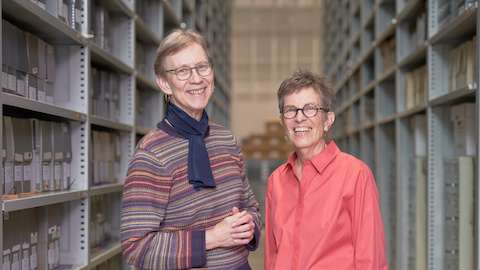 Barbara Loveland and Linda Powell standing in the massive stacks in the archives, facing the camera and smiling widely.