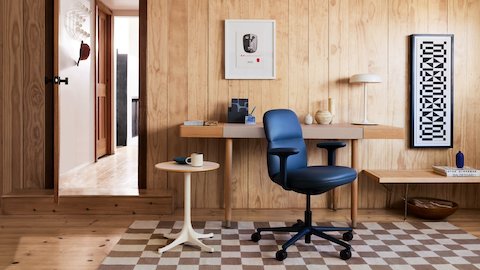 An upholstered dark blue leather office chair with arms sits at an angle, in front of a light wooden and tan leather desk, with minimal tchotchkes on the surface in neutral tones and dark blues. The walls are a light wood plank running vertically, and a small round white pedestal table sits below the desk with a coffee mug on top.