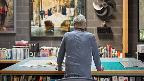 Don Chadwick, wearing a blue shirt, shown next to a table in his studio. The wall behind him features bookshelves filled with books and binders.