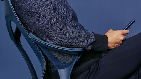 A man wearing a navy blue sweater sits in a Nightfall navy blue Cosm chair with leaf arms.