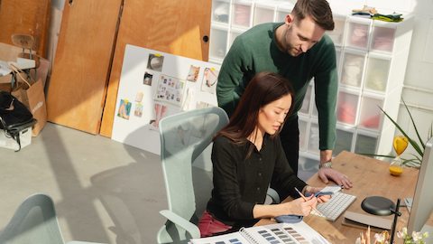 Two people collaborating at a desk in an office. Photos are pinned up on the wall behind them.