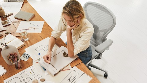 A woman sitting at a desk in her Aeron Chair, sketching shapes and designs on paper.