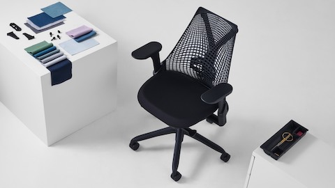 A black Sayl chair viewed from above, is positioned next to both a white cube, on top of which sits a folded arrangement of Revenio textile swatches and OE1 Trolly plastic clips, and a Tu Storage Pedestal with the top drawer open revealing a black Utility Tray made of ocean-bound plastic.