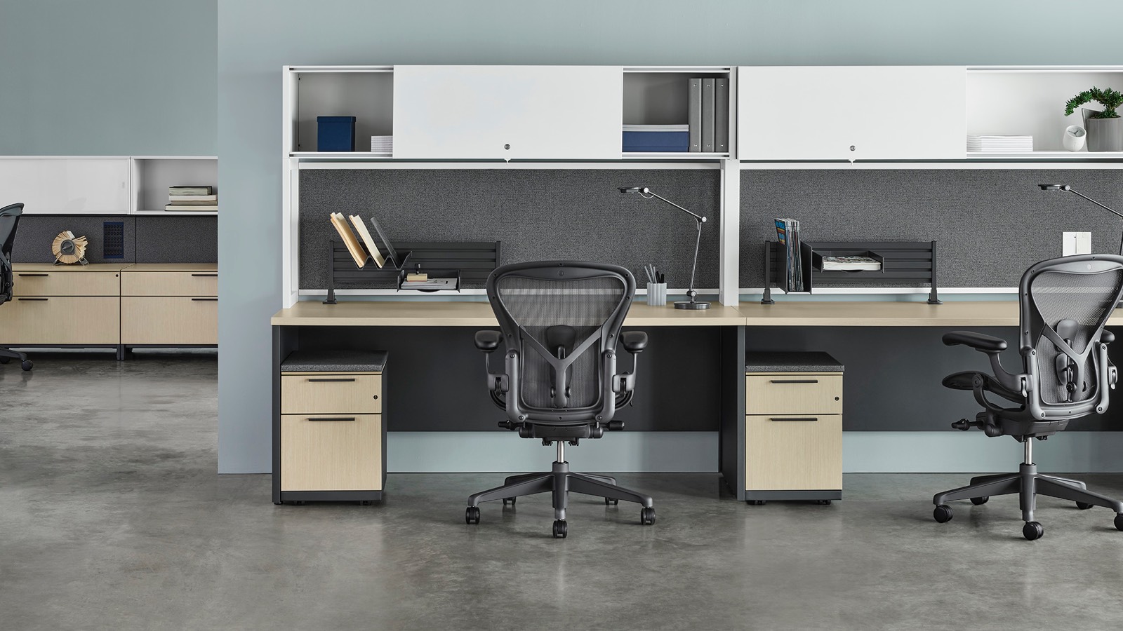 Three Canvas Metal Desks with white upper storage, gray fabric back panels, and light wood surfaces with another workstation in the background, all with dark grey Aeron chairs.