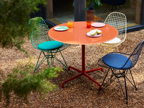 Herman Miller x HAY, escenario al aire libre con sillas Wire verdes, negras, azules y amarillas y una mesa Eames roja de hierro