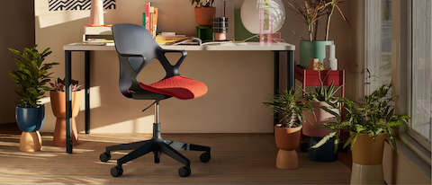 Uma cadeira Zeph escura com uma almofada de assento 3D Knit em vermelho vibrante em um home office ensolarado e cheio de plantas.