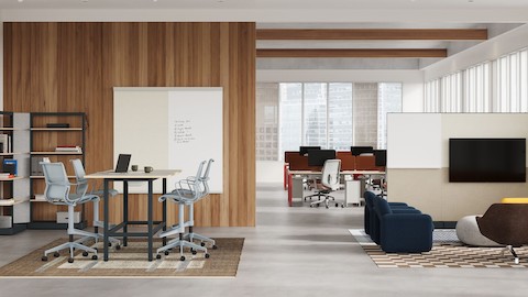 In an open floorplan office space with white walls and wood accents, featuring a standing table with gray Setu Stools around it, a lounge space in front of a screen, and rows of desks with computer monitors and gray Lino Chairs in the background.