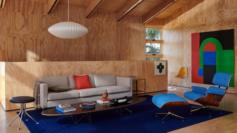 At home in a living room featuring all light tone woods, with an Eames Lounge & Ottoman in bright blue Ultramarine Checker, a gray Lispenard Sofa, a black Eames Elliptical Table, and a white Nelson Saucer Bubble Pendant, with colorful artwork and accessories nearby.
