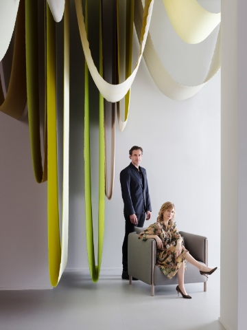 Designers Stefan Scholten (standing) and Carole Baijings (seated) pose next to hanging textiles in shades of green.