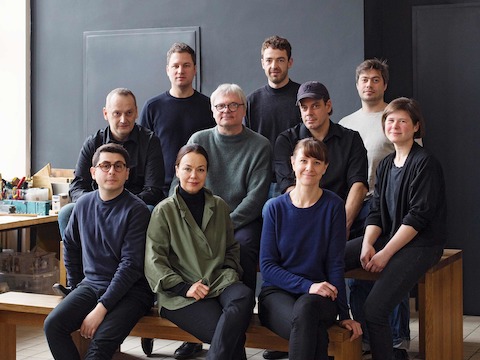 The men and women of Studio 7.5 sit grouped together on wooden benches in their workspace.