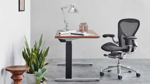 A black Aeron office chair shown with a sit-to-stand table and Eames Walnut Stool.