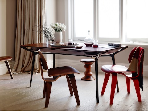 A medium woodgrain Carafe Table used for dining and surrounded by Eames Molded Plywood Chairs and an Eames Walnut Stool.
