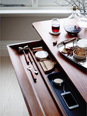 The main drawer of a medium wood Carafe Table, open to reveal a charging smartphone and other items inside.