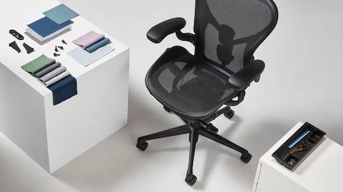 Una silla para oficina Aeron en negro vista desde arriba, posicionada junto a un cubo de exhibición blanco, sobre cuya superficie hay muestras textiles Revenio dobladas, sujetadores plásticos del carrito OE1 y una bandeja de utilidad de pedestal Tu de plástico oceánico recuperado.