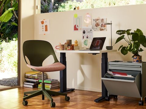 Una silla Zeph sin brazos en verde cerca de un escritorio de altura ajustable en blanco en una home office con luz solar.