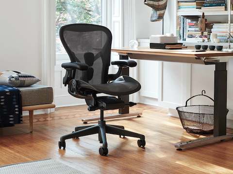家庭办公设置中，带木纹饰面的Renew坐姿-站姿切换两用办公桌的旁边有一张黑色的Aeron座椅。