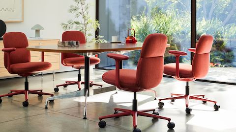 Quatre sièges Asari Herman Miller en rouge profond autour d'une table, dans une pièce éclairée par une fenêtre baignée de soleil.