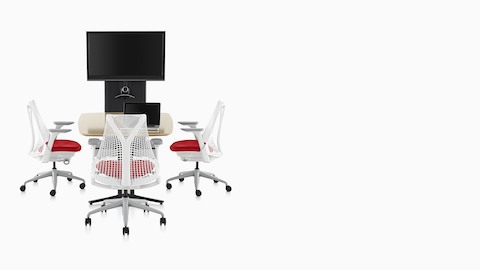 Three Sayl ergonomic office chairs with white backs and frames and red upholstered seats around a table facing an AV/VC One technology cart. Three Sayl ergonomic office chairs with white backs and frames and red upholstered seats around a table facing an AV/VC One technology cart.