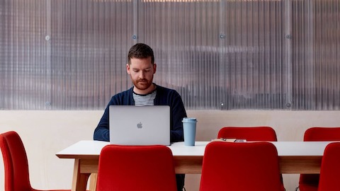 Ein Mann arbeitet mit einem Laptop an einem weißen Dalby Tisch mit 8 roten Viv Stühlen drum herum.