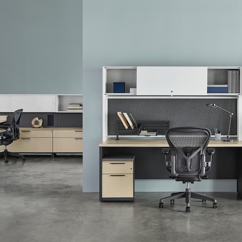 Mesa metálica Canvas com armário superior branco, painéis posteriores em tecido cinza, superfícies em madeira clara com outra estação de trabalho no fundo. Mesa metálica Canvas com armário superior branco, painéis posteriores em tecido cinza, superfícies em madeira clara com outra estação de trabalho no fundo.