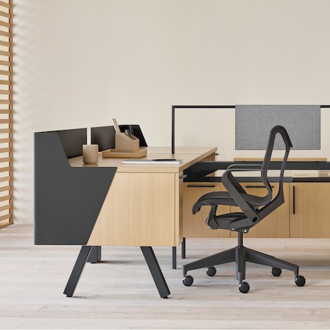 Una estación de trabajo Canvas Vista en madera clara y negro con pantalla de recato, panel de tela gris, y silla para oficinas Cosm negra.