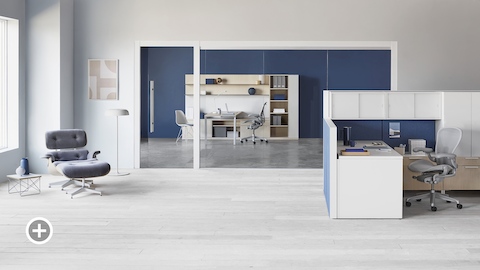 An office setting with a Canvas Wall workstation with blue panels and white overhead storage, a light gray Aeron office chair, a nearby Eames lounge chair in light gray, and a Canvas Private Office in the background. Select to go to the Individual Workstations page within the Canvas Lookbook.