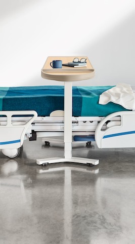 A patient room with a hospital bed covered with a blue and turquoise blanket and an overbed table with an ash finish top and white base.