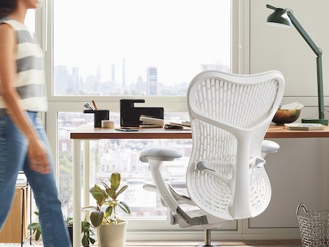 Una persona se acerca a una silla Mirra 2 en blanco, vista desde atrás, arrimada a un escritorio, con una ventada detrás.