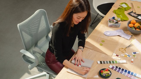 一个女人坐在办公桌前的Cosm座椅上，在记事本上涂写着。