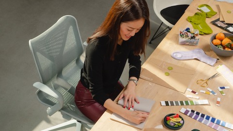 Mulher sentada à mesa em sua cadeira Cosm, escrevendo em um bloco de anotações.