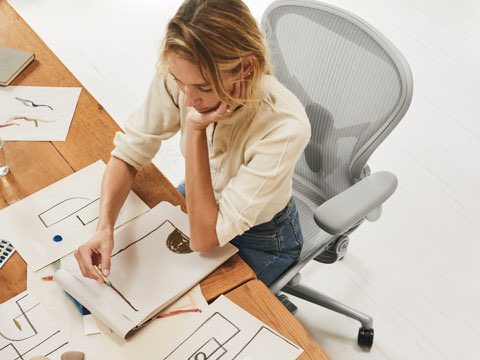 一个女人坐在办公桌前的Aeron座椅上，在纸上勾画着一些形状和设计。