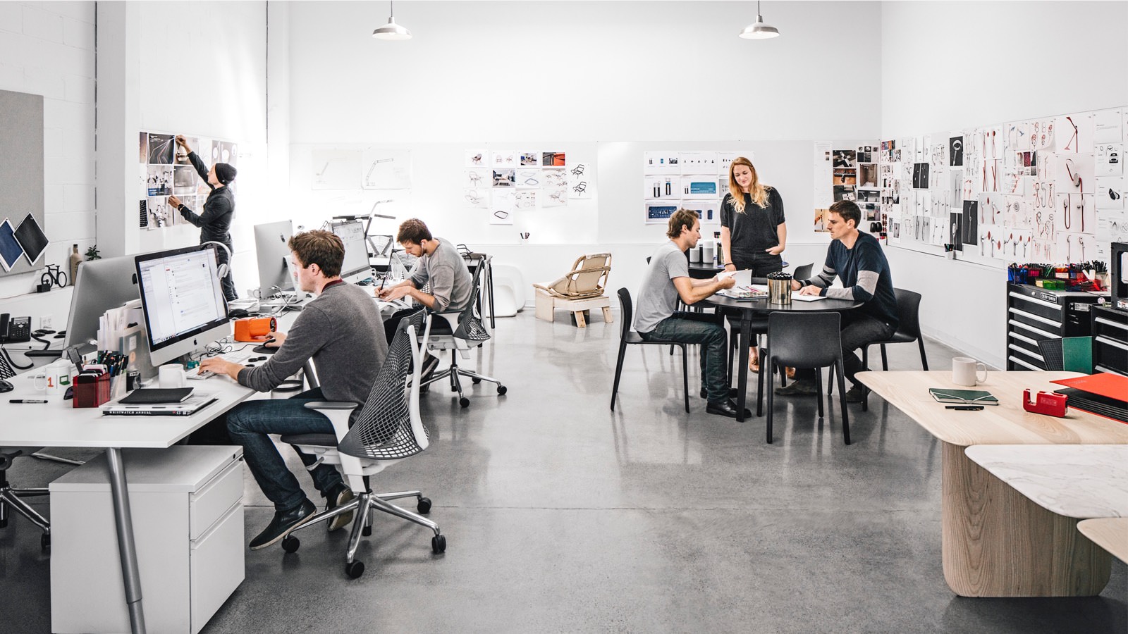 A photo of a workplace with people working at computers while sitting at long desks in Aeron Chairs. A trio of colleagues meet nearby.