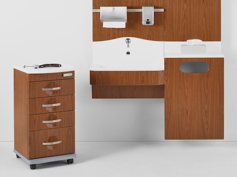 A Compass casework wall in a medium walnut finish and a white solid surface sink and surface with a Compass supply cart next to it.