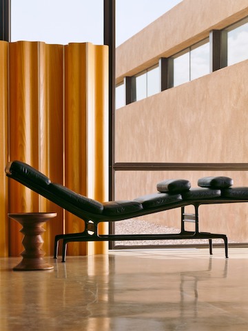 Profile view of a black leather Eames Chaise next to an Eames Walnut Stool in an office building. 