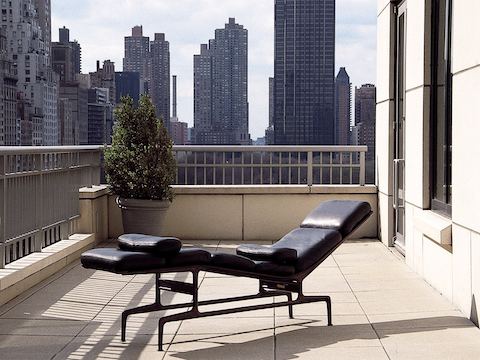 Un cuero negro Eames Chaise situado en un balcón con vistas a un horizonte urbano.