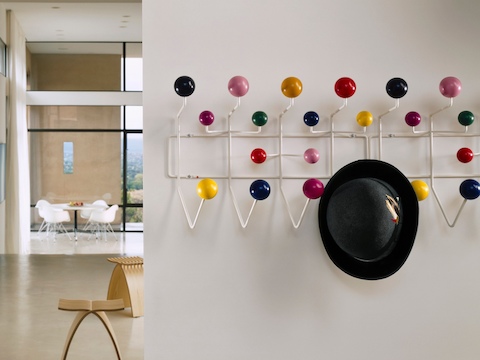 A man's hat hangs from one of two adjacent Eames Hang-It-All storage racks, both with a white wire frame and multicolored knobs.