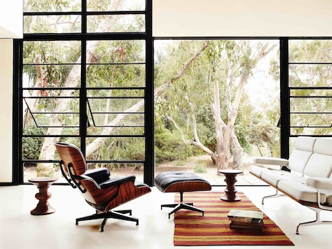 Una silla de salón Eames de cuero negro y una otomana, un sofá Eames y dos taburetes de nogal Eames cerca de una pared de vidrio.