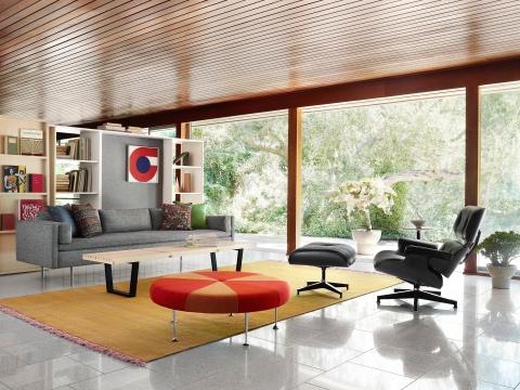 Um couro preto Eames Lounge Chair e otomano e um cinza Bolster Sofa ancorar um ambiente residencial com uma vista exterior.